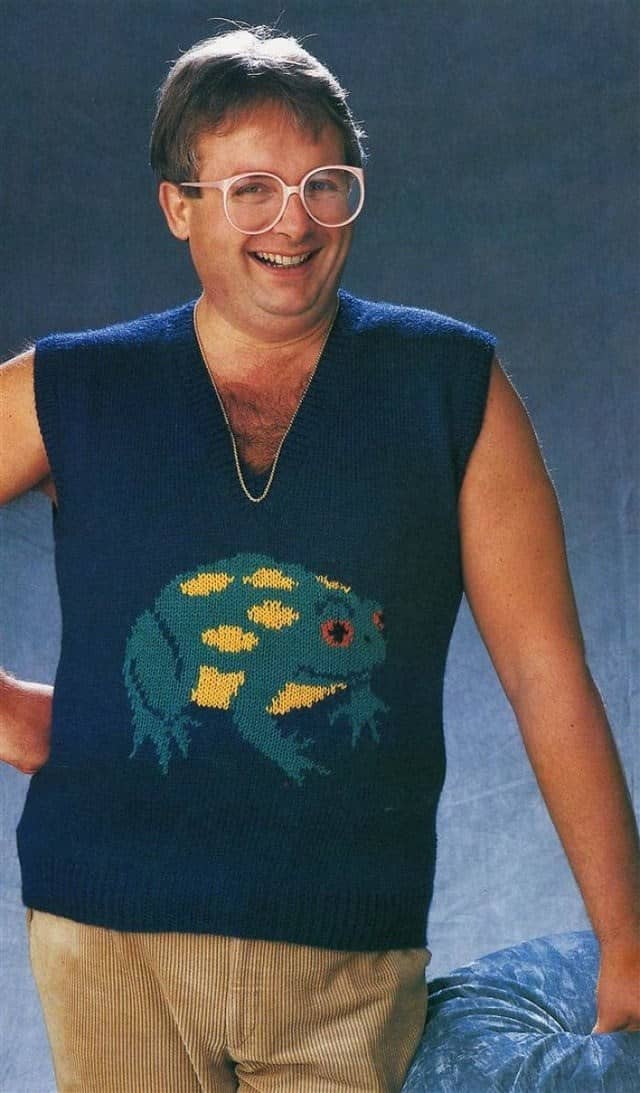 A man wearing a navy sweater vest with a large knitted frog graphic and oversized round glasses.