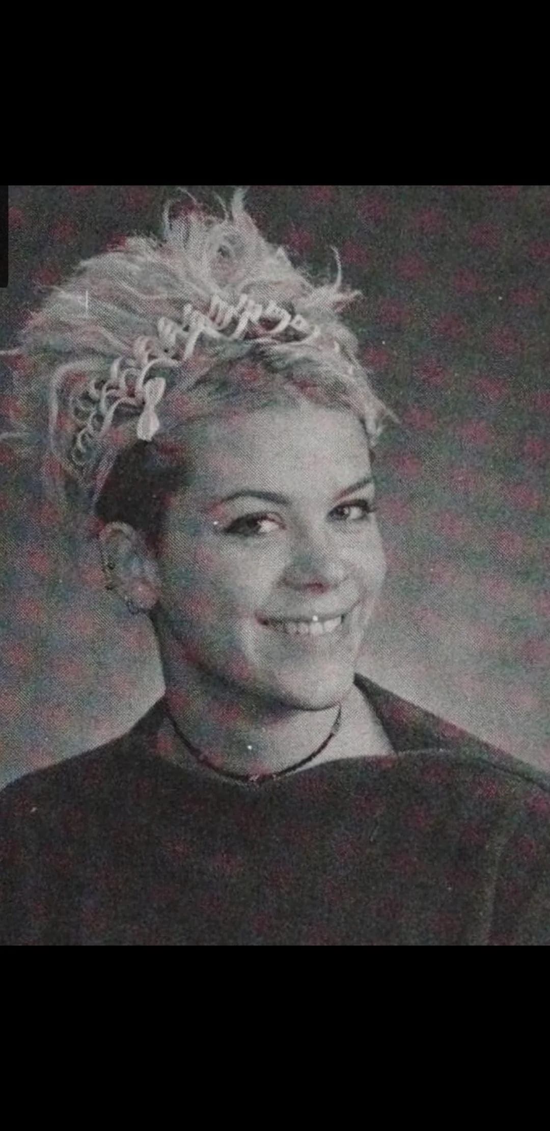 Grainy yearbook headshot of a teen with a spiky updo packed with tiny clips, a choker necklace, and a sly smile.