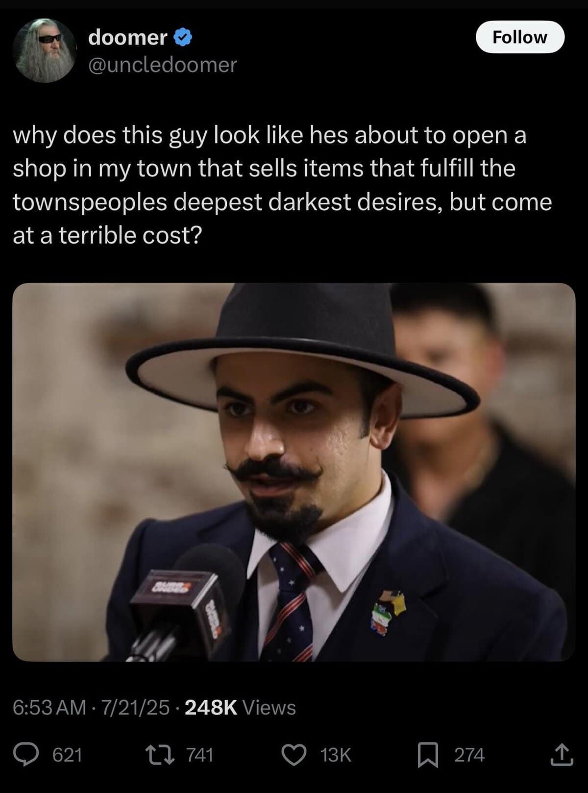 man in a dramatic wide-brimmed hat and curled mustache being interviewed; tweet suggests he looks like a mysterious shopkeeper from a fantasy town.