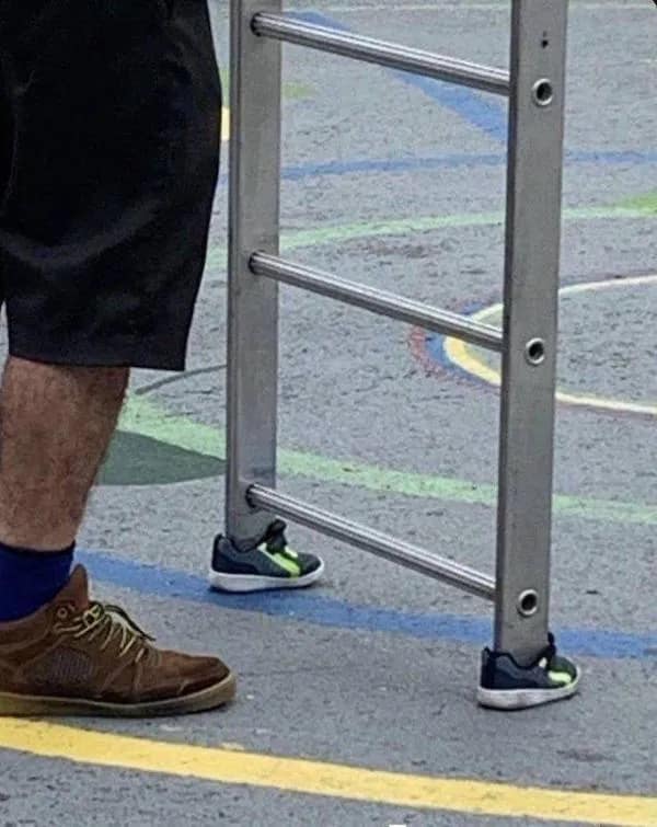 Aluminum ladder feet fitted with tiny toddler sneakers on pavement next to a worker’s shoe.