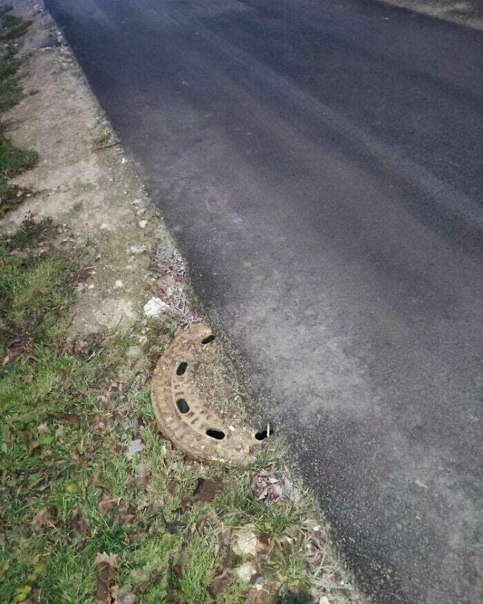 A broken, half-circle manhole cover on the shoulder of a paved road, leaving a large and dangerous semi-circular hole completely exposed.