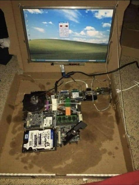A deconstructed laptop computer that has been rebuilt inside of a greasy and stained cardboard pizza box, running the Windows XP operating system.