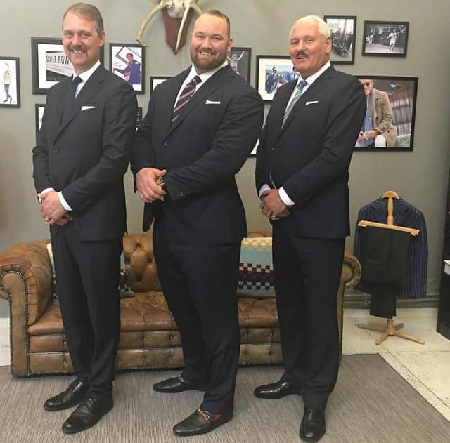 An absolute unit of a man in a suit standing between two normal-sized men, appearing twice as wide