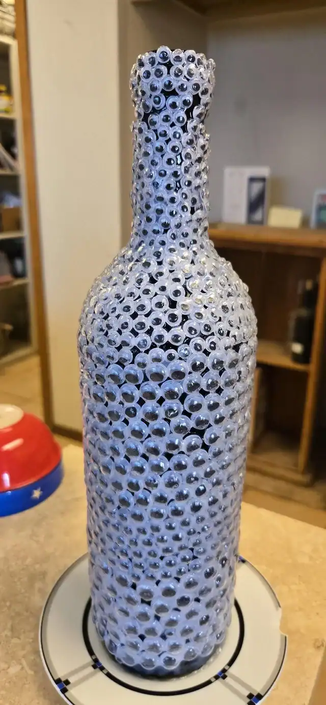 Wine bottle completely covered in hundreds of plastic googly eyes eyebombing art.
