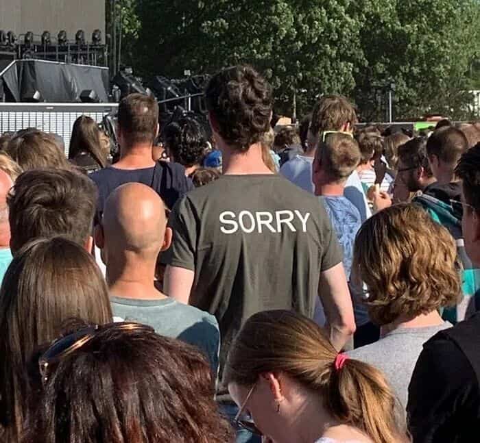 A tall people problems photo of a man at a concert wearing a shirt that says "SORRY" to the people behind him.