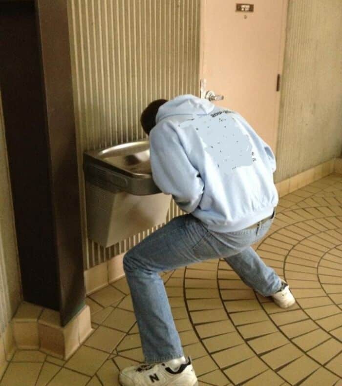 A tall people problems photo of a man doing the splits just to reach a low water fountain.