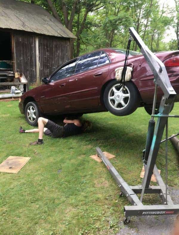 A classic osha meme featuring a mechanic working under a car that is suspended in the air by a single strap looped through the rear windows and attached to an engine hoist.