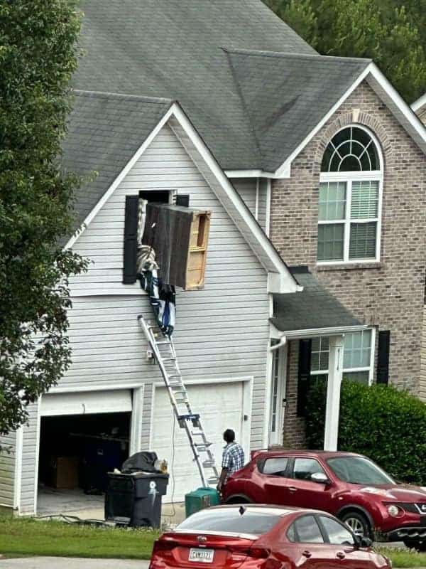 A chaotic osha meme showing movers attempting to slide a massive piece of furniture out of a second-story window using a single ladder as a makeshift slide.