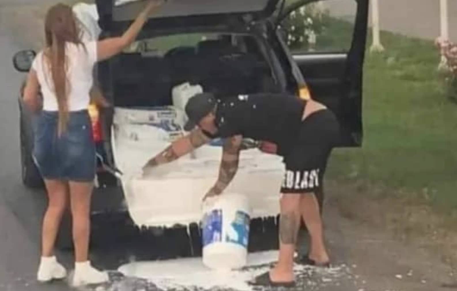 Man frantically cleaning spilled white paint from the trunk of a black car after bucket tipped.