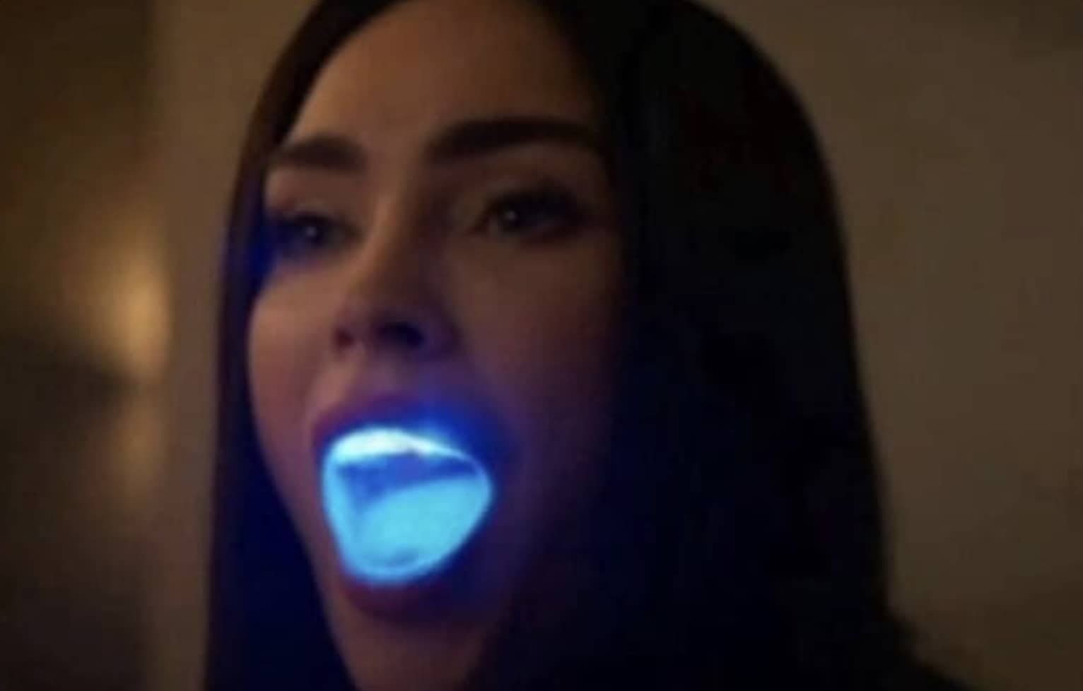 A woman with a glowing blue light inside her open mouth in a dark setting.