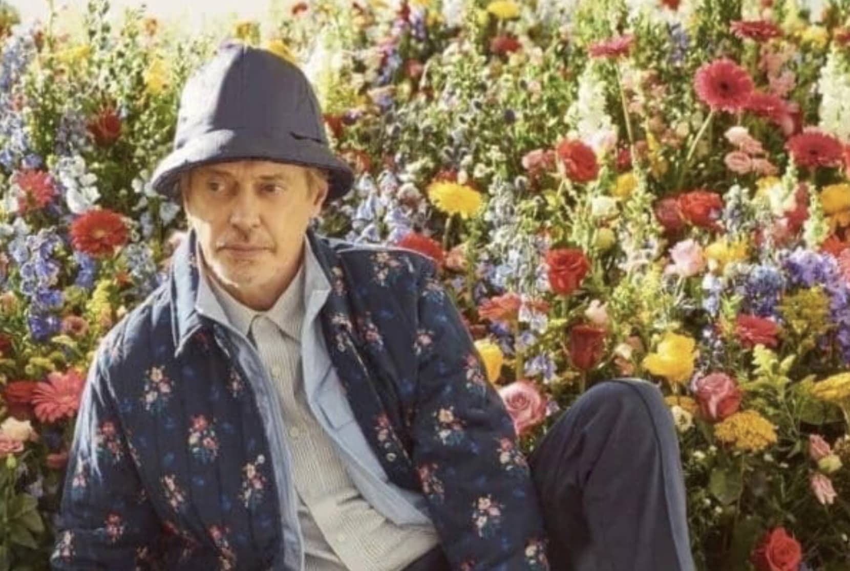 Actor Steve Buscemi wearing a floral jacket and blue hat sitting in front of flowers.