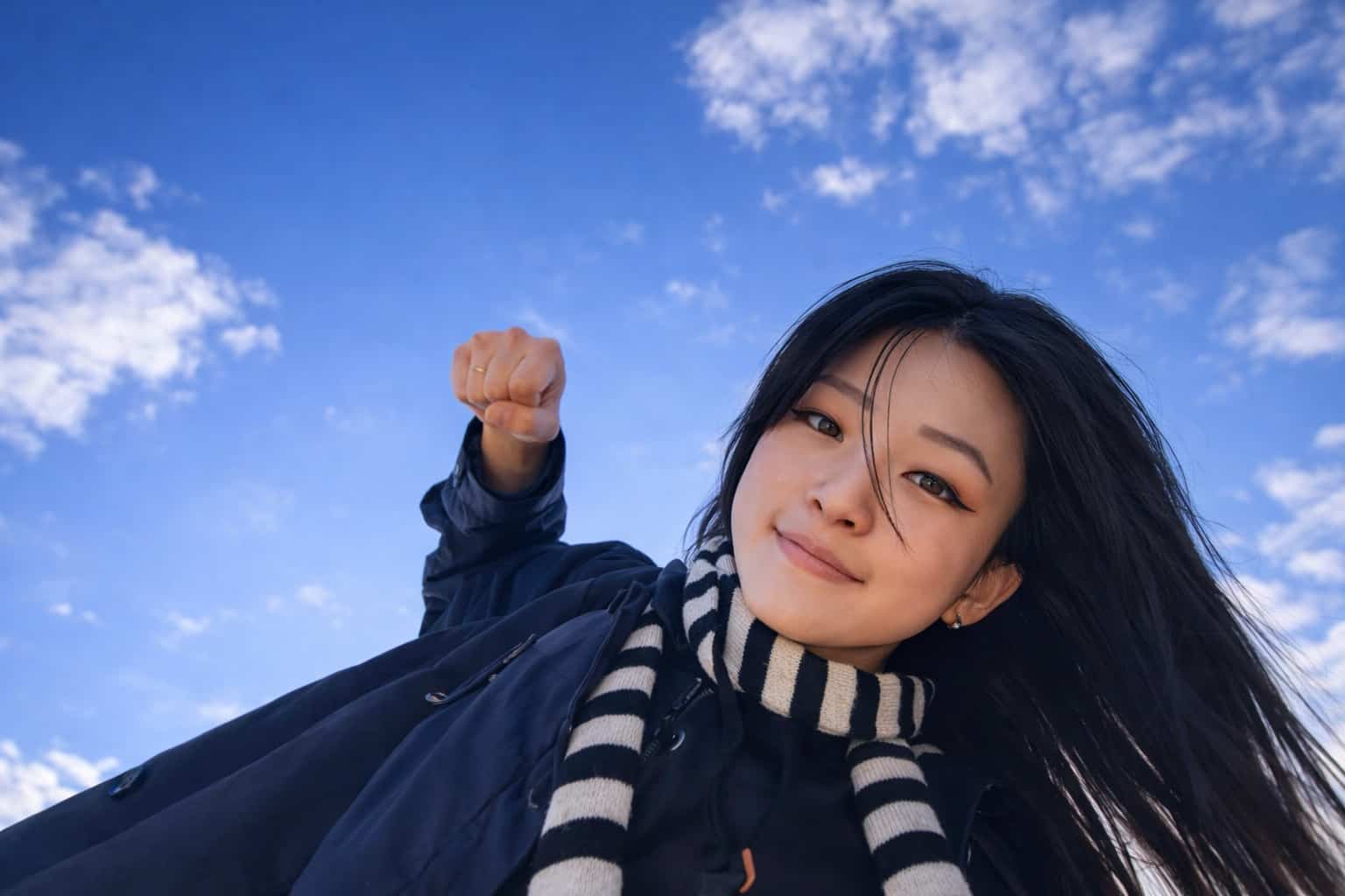 Young woman in a scarf posing with a raised fist against a blue sky for relatable memes.