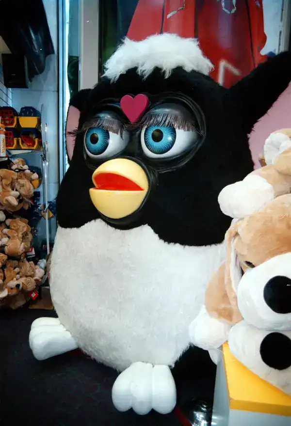 nostalgic 90s picture showing a giant, slightly terrifying Furby display model in a toy store, featuring its signature large blue eyes, yellow beak, and black and white fur