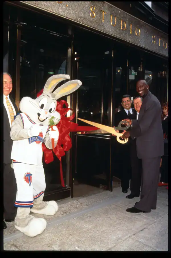 iconic promotional moment in 90s pictures showing Michael Jordan standing next to a person in a full Bugs Bunny mascot costume, both holding giant scissors to cut a red ribbon for a store opening.