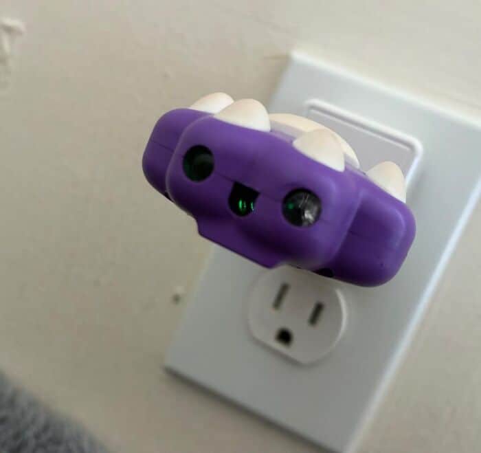 cute example of inanimate objects with faces featuring a purple plug-in device in a wall outlet, using its green indicator lights and plastic mold to create a happy, adorable little monster face.