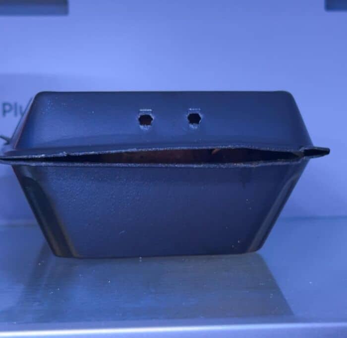 slightly menacing entry of inanimate objects with faces showing a black takeout container slightly cracked open like a wide mouth, with two ventilation holes acting as tiny eyes staring back from the fridge.