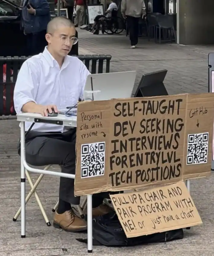 gritty programmer meme depicting a self-taught developer sitting at a folding table on a city sidewalk with a laptop and a cardboard sign. The sign reads: "SELF-TAUGHT DEV SEEKING INTERVIEWS FOR ENTRY LVL TECH POSITIONS. PULL UP A CHAIR AND PAIR PROGRAM WITH ME!"