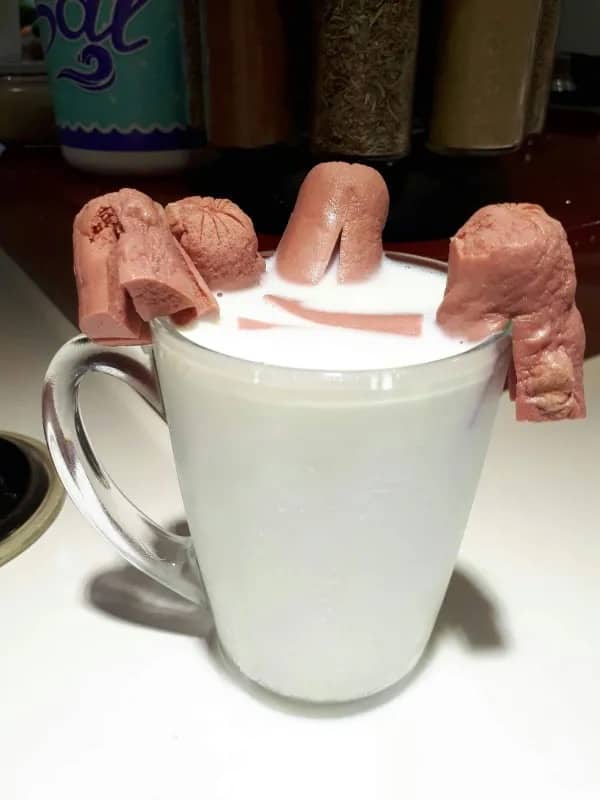 Disgusting food image of cut hot dogs placed on the rim of a mug filled with milk.