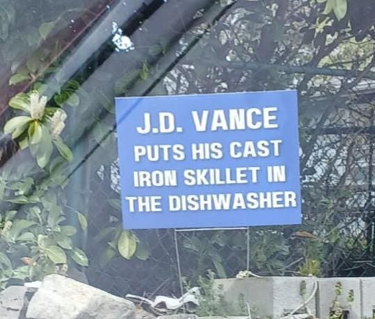 A photo of a blue political-style yard sign nestled in some greenery. The white block lettering delivers a devastating non-political insult: "J.D. VANCE PUTS HIS CAST IRON SKILLET IN THE DISHWASHER," attacking his domestic credibility.