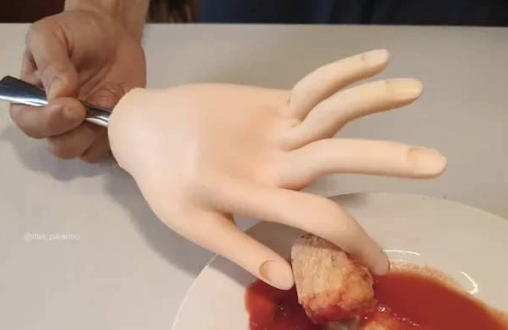 Person using small prosthetic hand attached to fork to dip food into red sauce on plate.