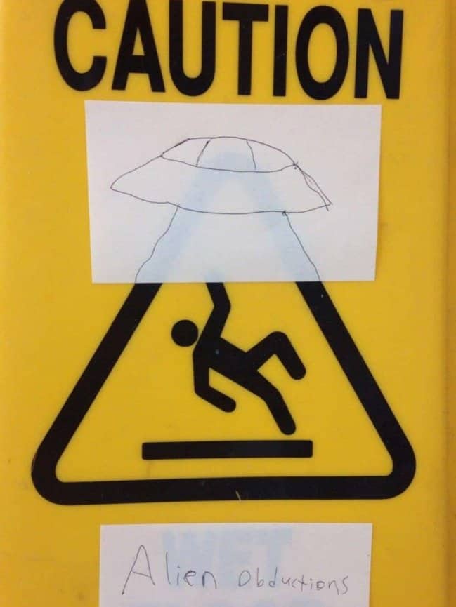 A wet floor sign modified with paper cutouts to depict an alien abduction.