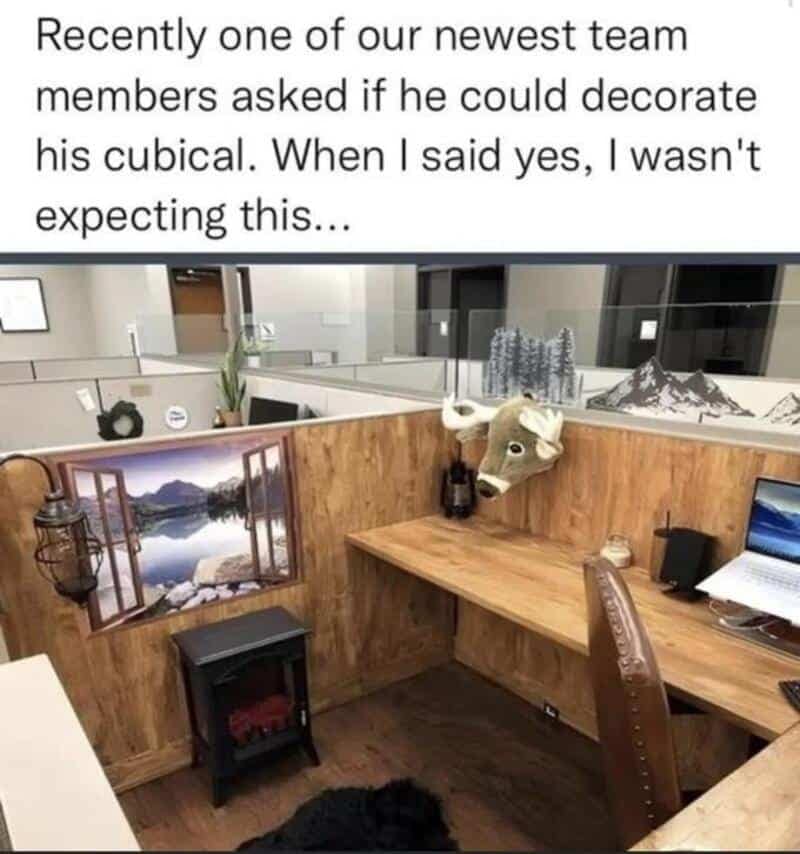 An office cubicle transformed into a rustic hunting lodge, complete with wood paneling, a mounted deer head, a lantern, and a miniature electric fireplace.