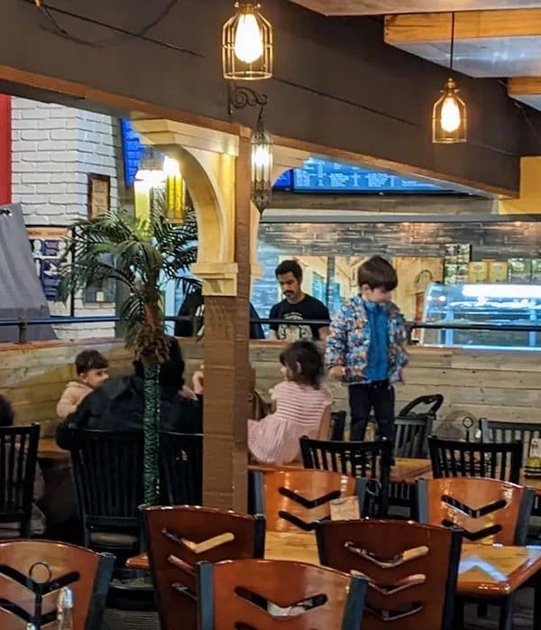 Young boy standing on top of a restaurant table while other diners sit nearby.