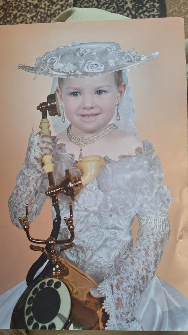 Little girl in a lace gown, flower hat and pearls posing with an antique phone.