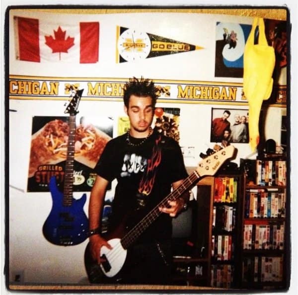 Spiky-haired teen playing bass in a bedroom full of Michigan banners and VHS tapes.