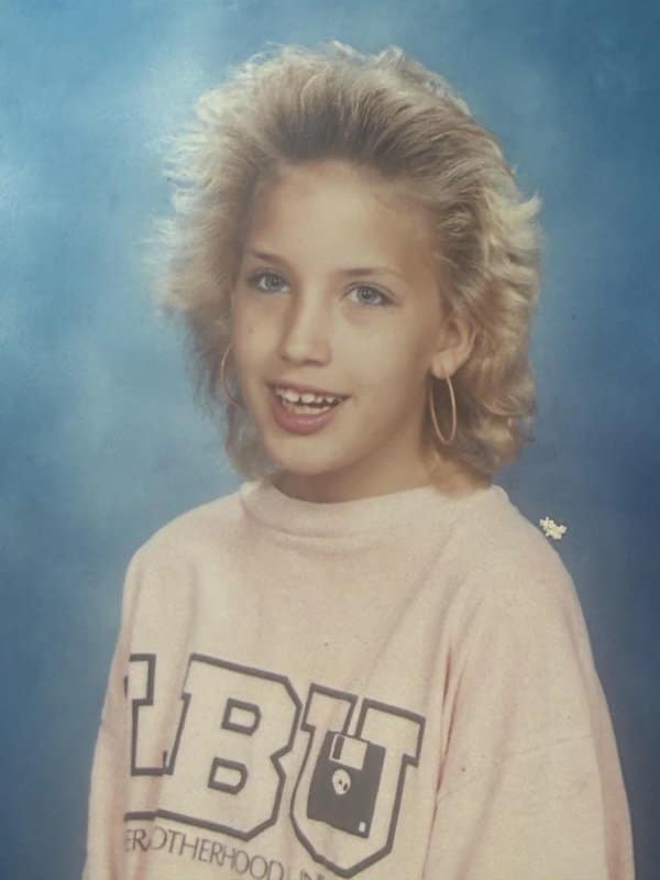 Girl with huge feathered hair, hoop earrings and a pink sweatshirt on a blue backdrop.