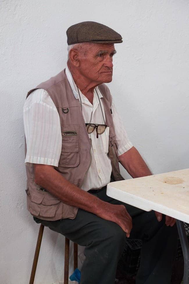 Serious older man in a flat cap and utility vest seated at a white plastic table