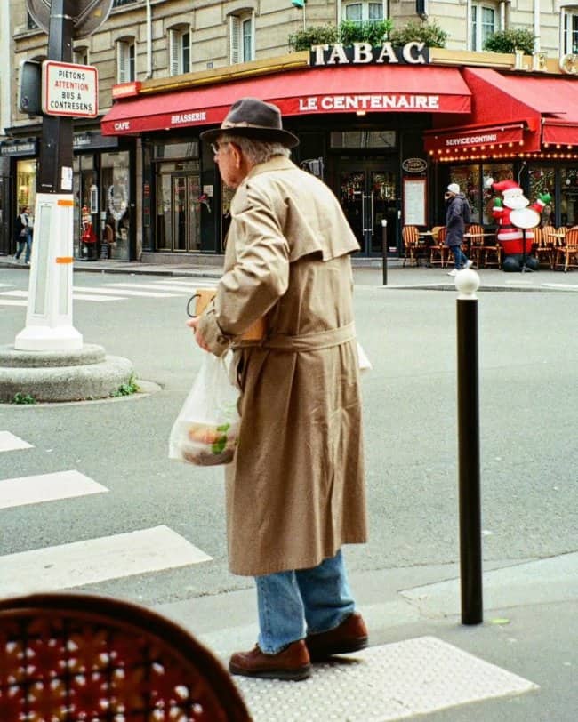 Cool grandpa in a trench coat and fedora carrying groceries across a Paris street
