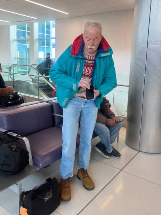 Grandpa in a teal jacket and patterned sweater checking his phone in an airport