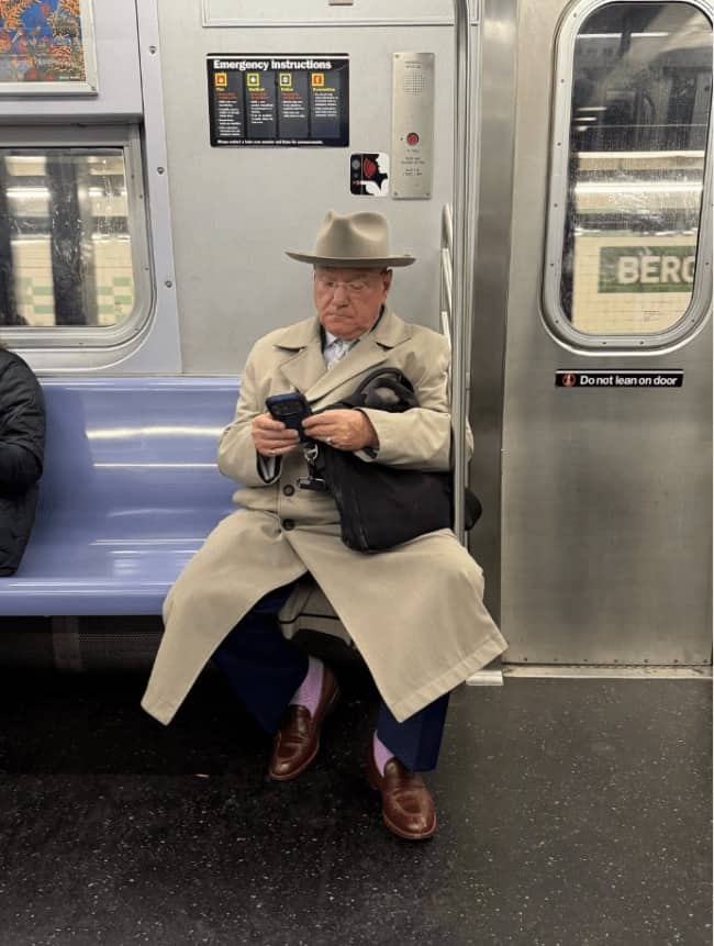 Older man in a fedora, trench coat, and pink socks riding the subway