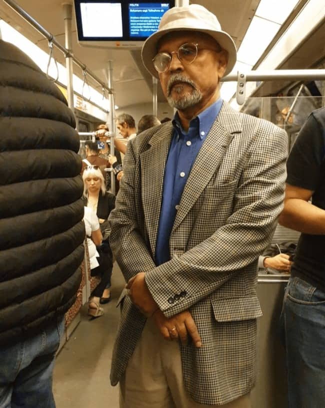 Older man in a bucket hat, round glasses, and houndstooth blazer standing on a train