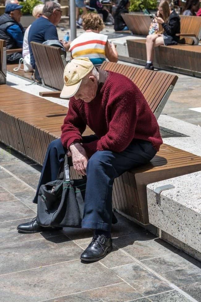 Older man in a burgundy sweater, cap, and polished shoes resting on a city bench