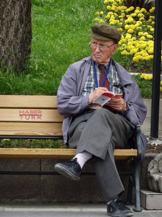 Older man in a flat cap, plaid shirt, and tie reading a book on a park bench