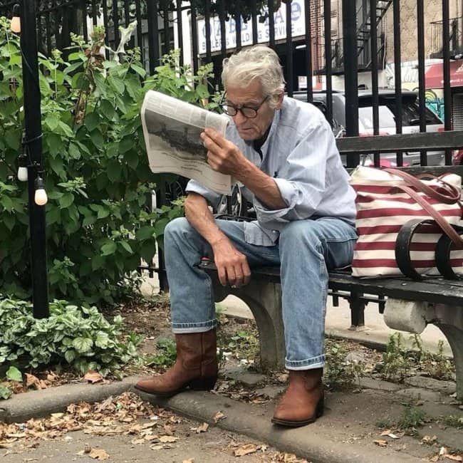 Silver-haired grandpa in jeans and cowboy boots reading a newspaper on a park bench