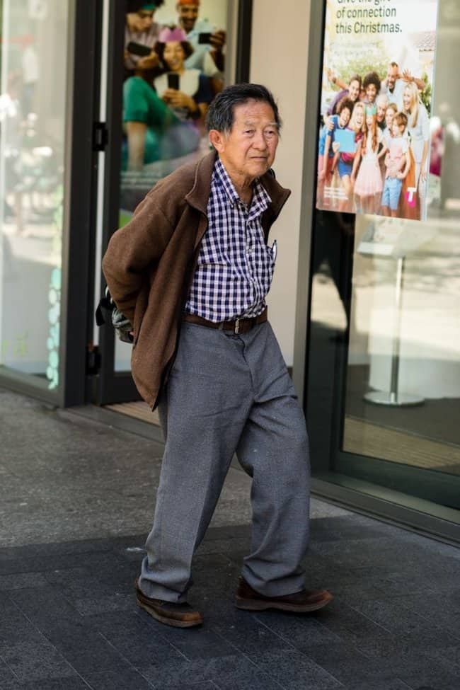 Older man in a brown jacket, checked shirt, and grey slacks walking out of a store