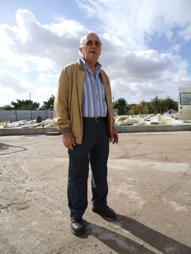 Older man in a tan bomber jacket and striped shirt standing on a dirt lot