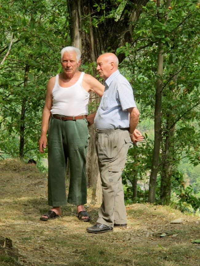 Two older men chatting on a forest trail, one in a white tank top and sandals