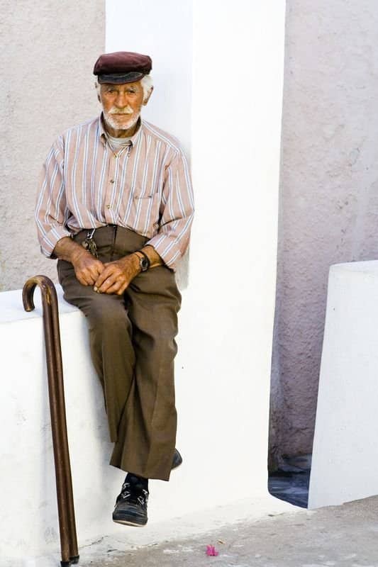 Older man in a burgundy cap and striped shirt seated on a white wall beside his cane