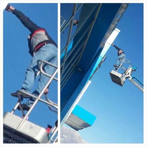 Worker standing on the guardrail of a lift basket, stretching to reach a building's overhang.