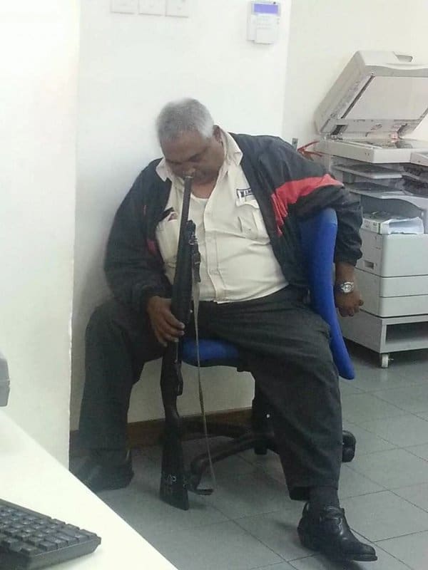 Security guard dozing in a blue office chair beside a copier, holding a rifle upright.