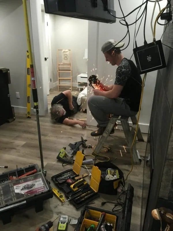 Man on a stepladder spraying angle grinder sparks over a coworker lying on the floor.