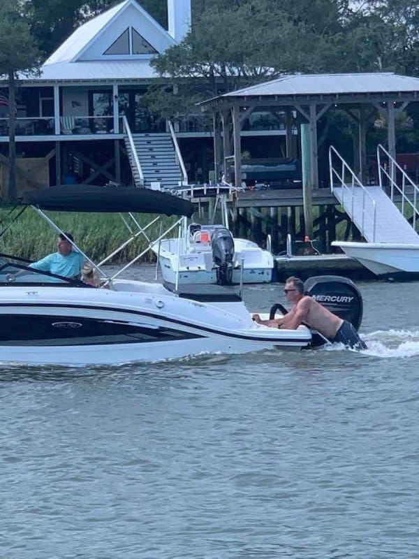 Shirtless man clinging to the back of a moving motorboat as it pulls away from a dock.