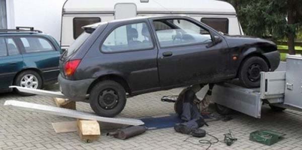 Small black car teetering off a trailer on wooden blocks while a man works underneath.
