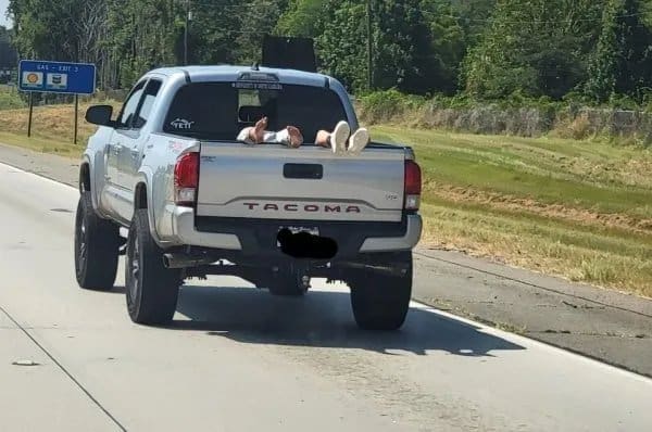 Passenger lying in a pickup truck bed on the highway, feet propped on the tailgate.