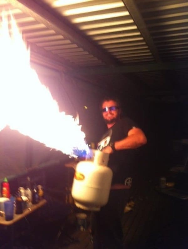 Grinning man in sunglasses shooting a huge flame from a propane tank under a patio roof.