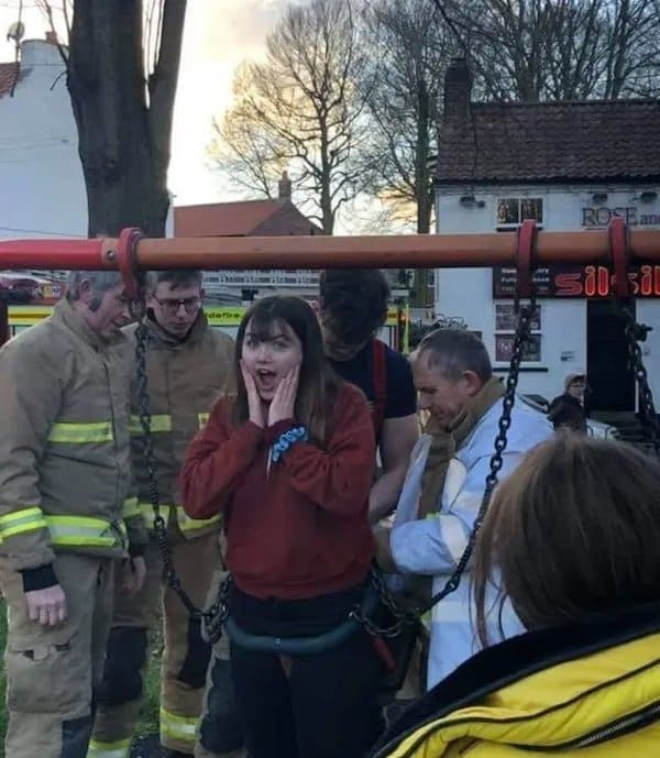 Young woman stuck in a toddler swing covering her face as firefighters work to free her.